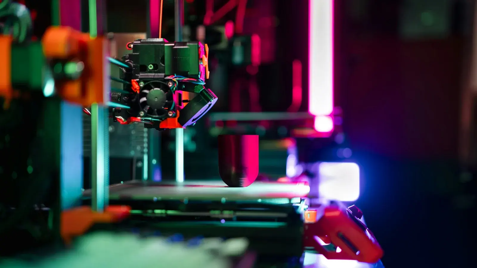 Symbolbild: Der Druckkopf eines 3D-Druckers arbeitet über einem dunklen zylindrischen Werkstück; farbige Lichtstreifen leuchten im unscharfen technischen Hintergrund. Das Bild zeigt weder Planitz noch das angekündigte Makerfestival.