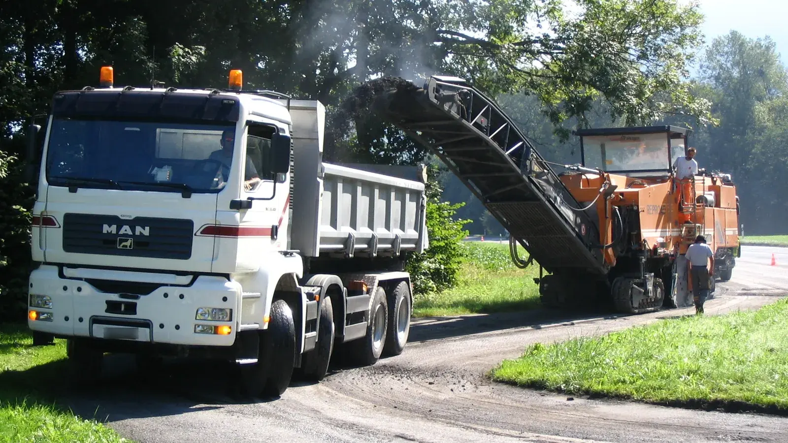 Archiv- und Symbolbild von 2005: Eine orangefarbene Straßenfräse befördert abgetragenen Asphalt über ein Förderband in einen weißen Lastwagen; zwei Arbeiter stehen an beziehungsweise auf der Maschine.