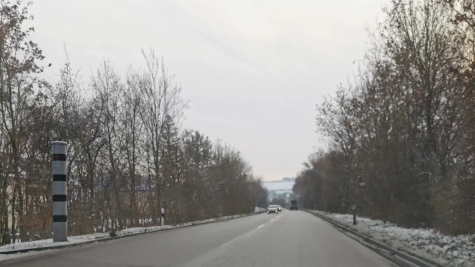 Symbolbild vom 15. Dezember 2022: Eine schwarz-weiß gestreifte Blitzersäule steht links neben einer verschneiten, von Bäumen gesäumten Straße in Bad Mergentheim; in der Ferne fahren mehrere Fahrzeuge.
