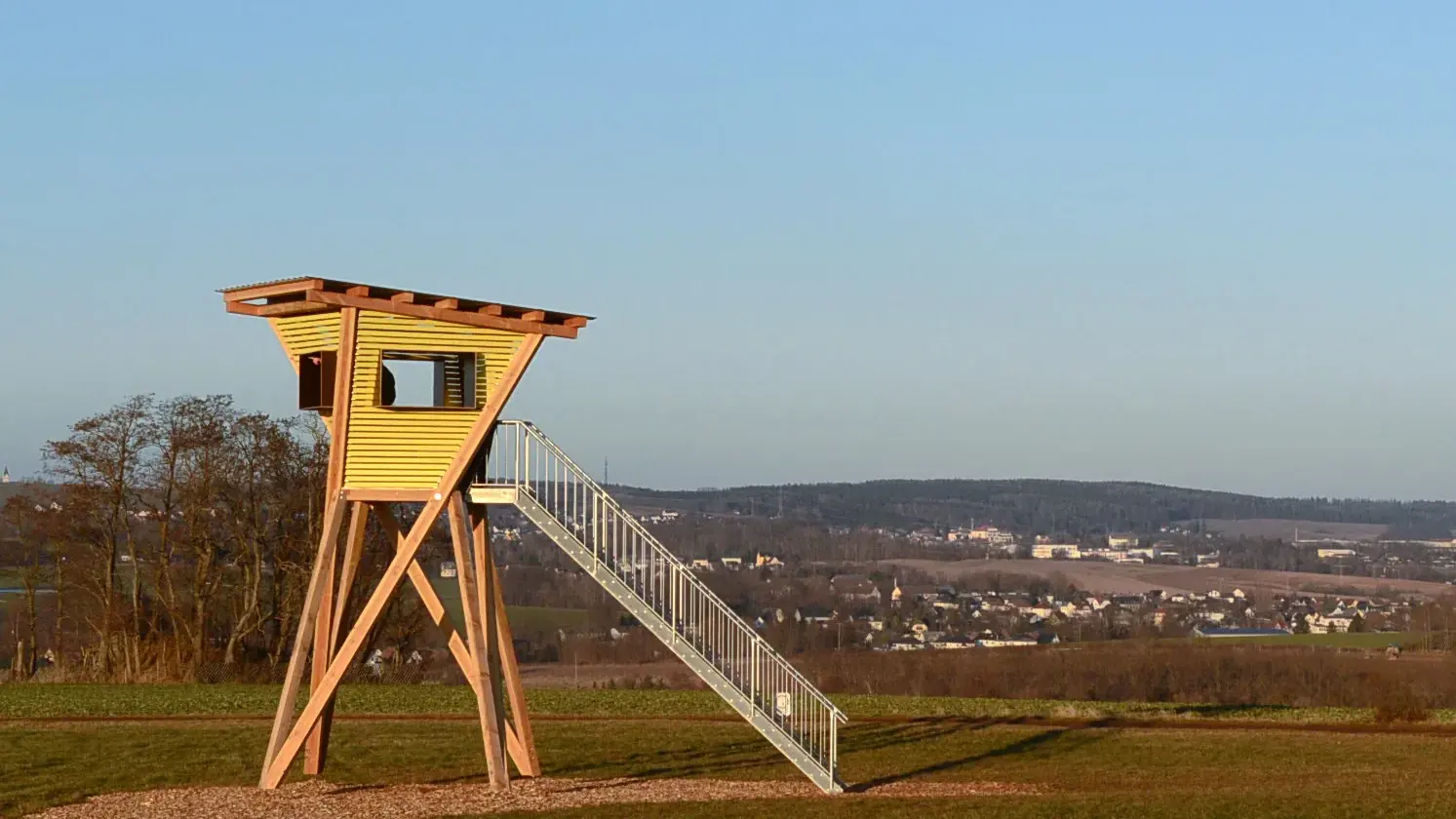 Archivaufnahme vom Januar 2026: Das hohe hölzerne Kunstwerk „Twisted Peek“ mit Außentreppe steht auf einer Wiese vor Feldern und der Oberlungwitzer Ortsansicht.