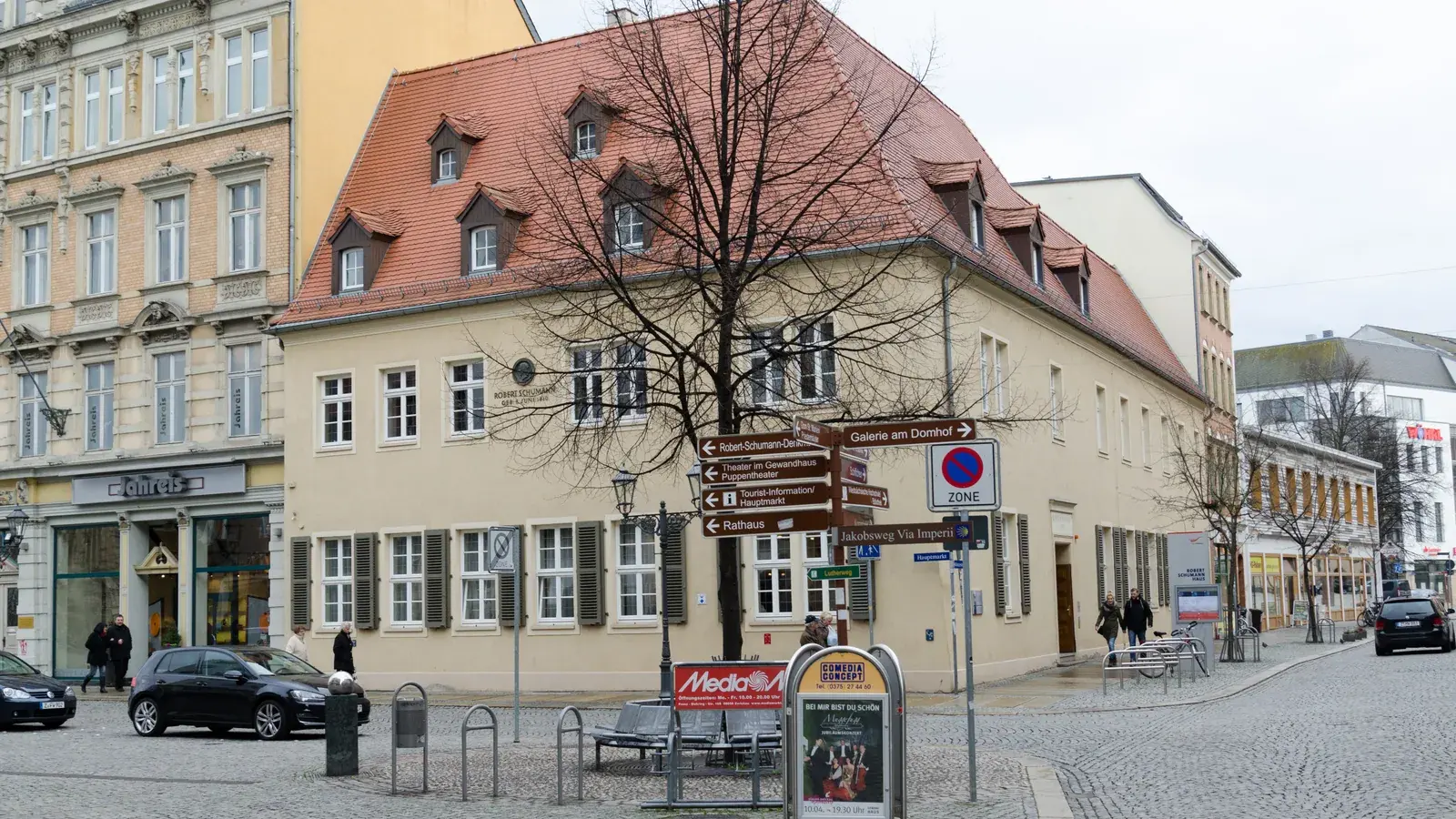 Das beige Robert-Schumann-Haus mit rotem Dach am Zwickauer Hauptmarkt, davor ein kahler Baum, Wegweiser und Straßenmobiliar.