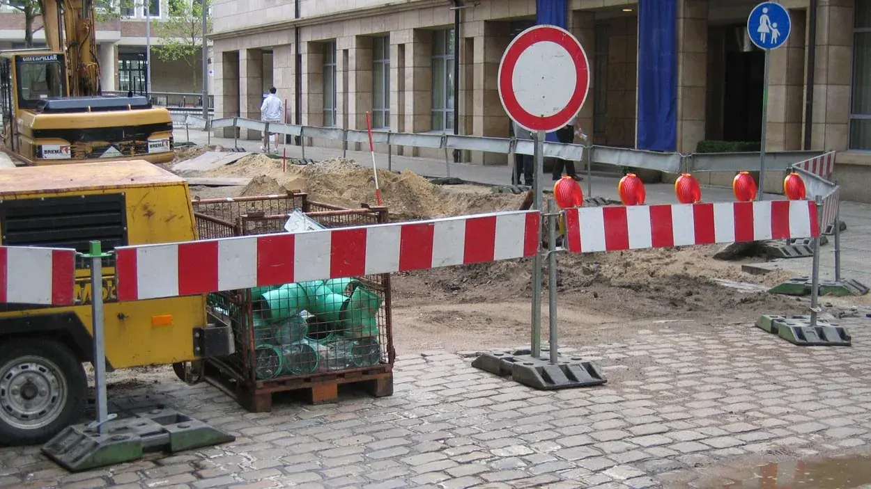 Symbolbild: Rot-weiße Absperrschranken und ein rundes Durchfahrtsverbot stehen vor einer aufgerissenen gepflasterten Baustellenfläche.