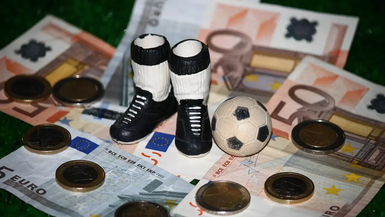 Eine kleine Fußballfigur mit Ball steht zwischen Eurobanknoten und Münzen auf grünem Untergrund.