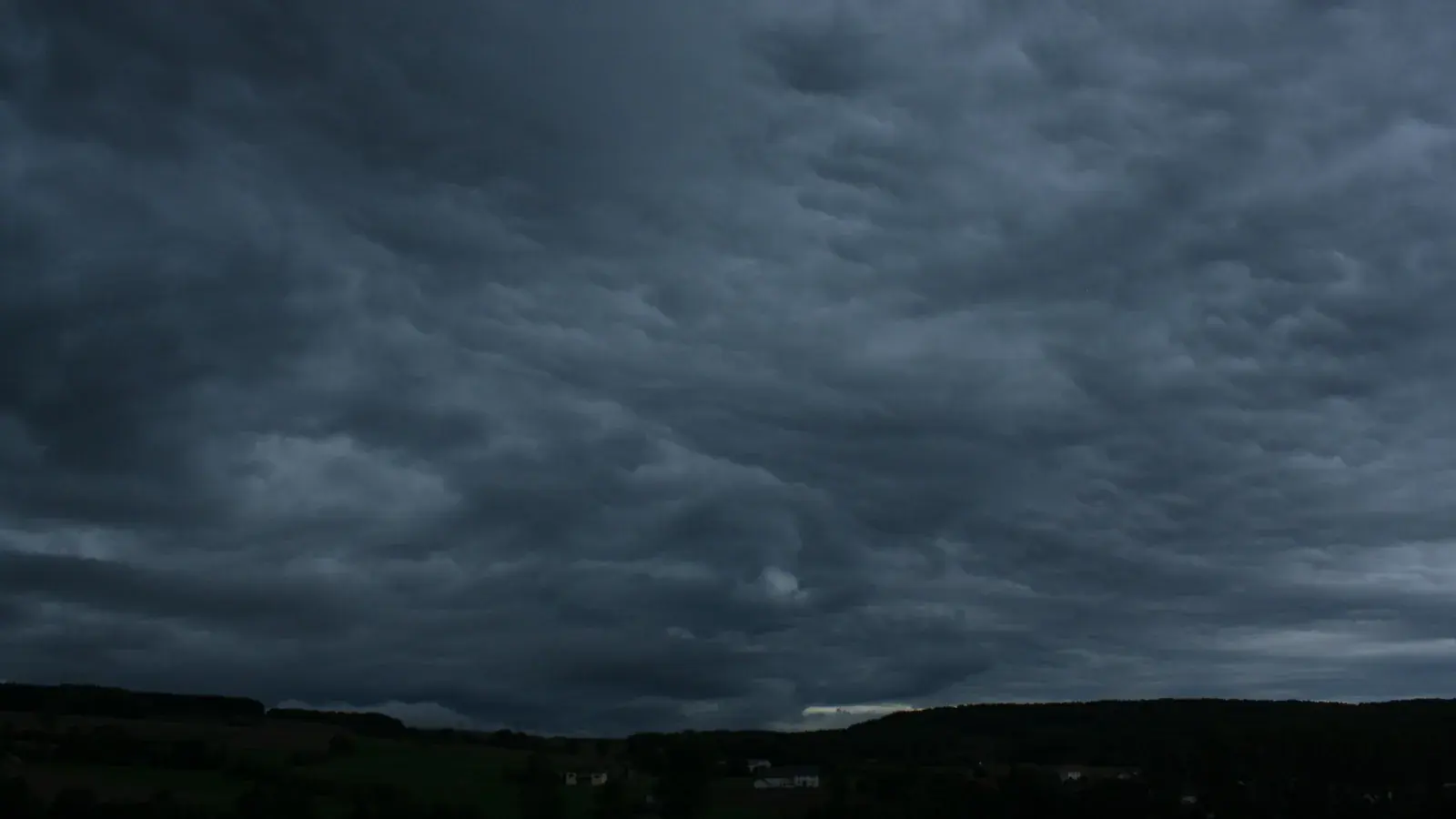 Symbolbild: Eine dunkle, stark strukturierte Wolkendecke liegt über einer kaum erkennbaren ländlichen Hügellandschaft.