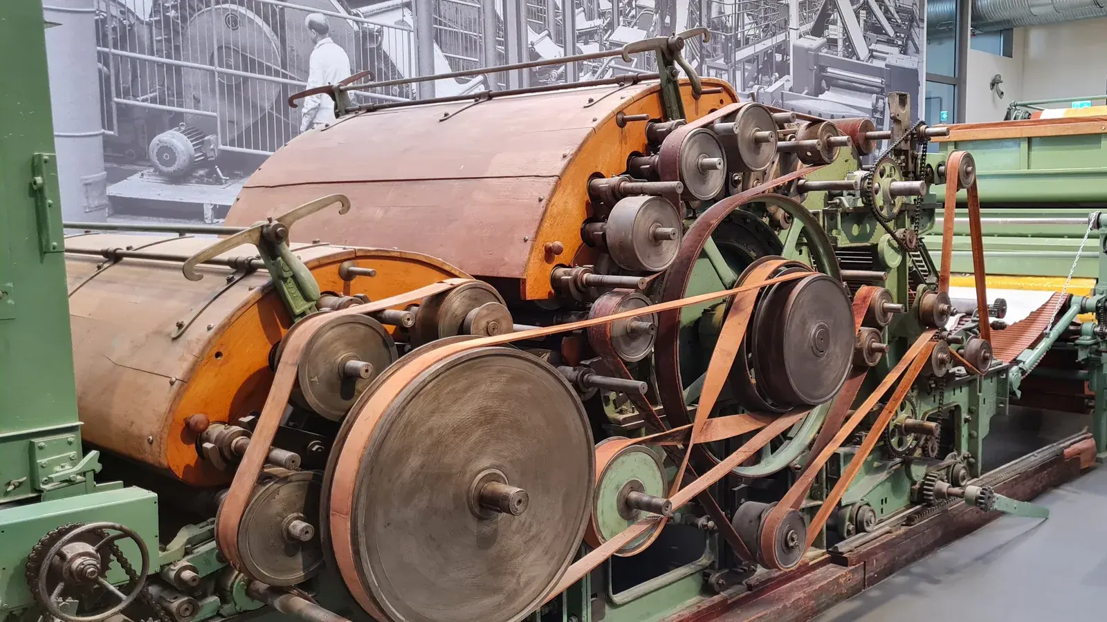 Archivaufnahme von 2023: Eine grün und orange lackierte historische Fertigungsanlage mit zahlreichen Walzen, Riemen, Ketten und großen Scheiben steht vor einem schwarz-weißen Fabrikfoto.