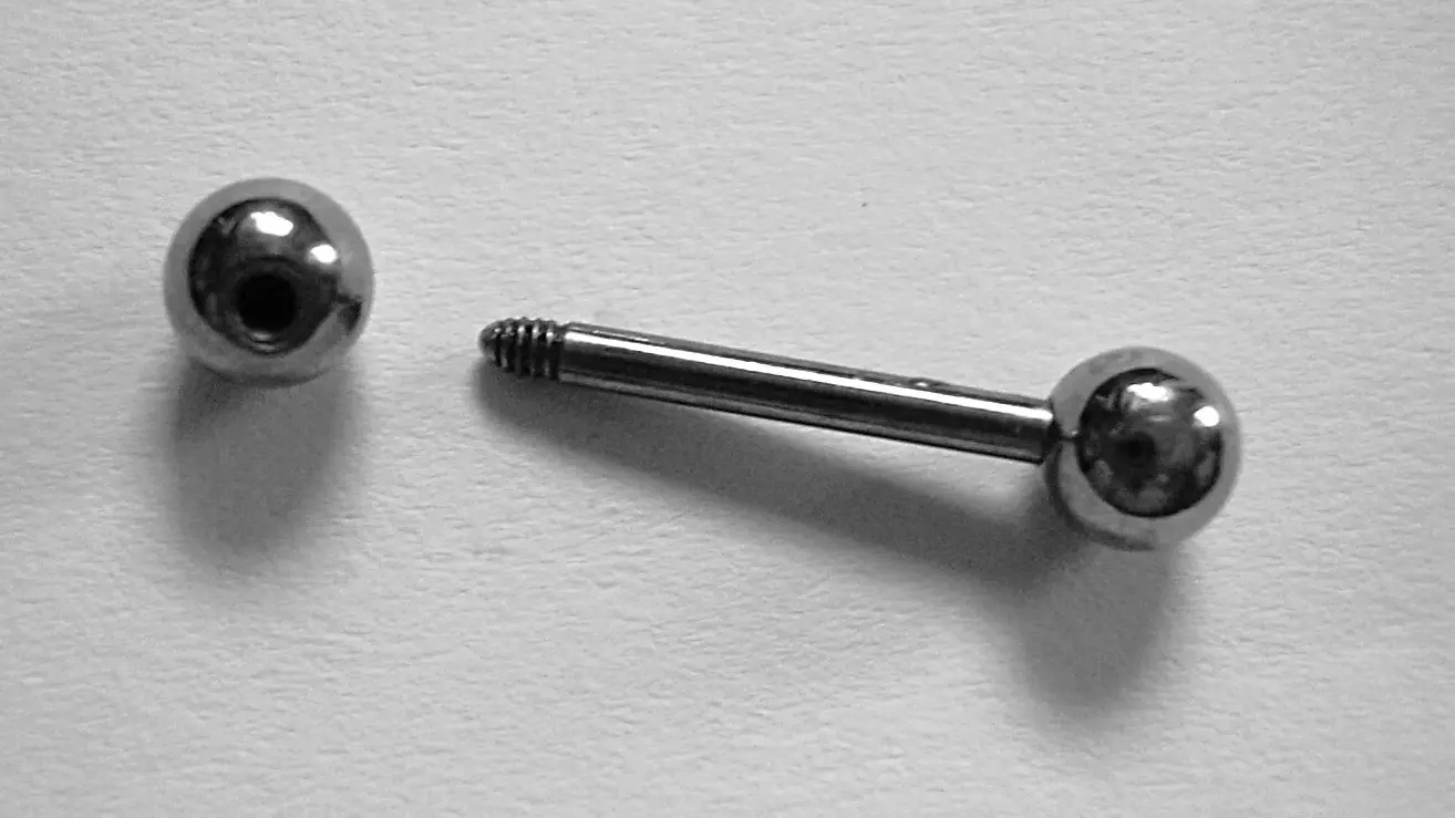 Symbolbild: Ein silberfarbener Piercing-Stab mit Schraubkugel liegt neben einer zweiten Metallkugel auf hellem Untergrund.